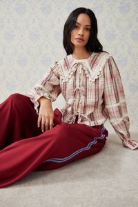Pantaloni larghi rossi con strisce laterali blu abbinati a una blusa rosa a quadri con rifiniture in pizzo e chiusura a fiocco, posizionati su uno sfondo a motivo.