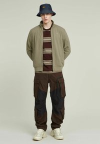 Giacca zip-up verde oliva sopra una camicia marrone a righe, abbinata a pantaloni cargo marrone scuro con accenti neri e sneakers beige.