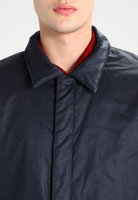 Dunkelblauer Polyester-Mantel mit großem Kragen, der eine glatte Textur und einen subtilen Glanz aufweist. Unter dem Kragen sind rote Details sichtbar.