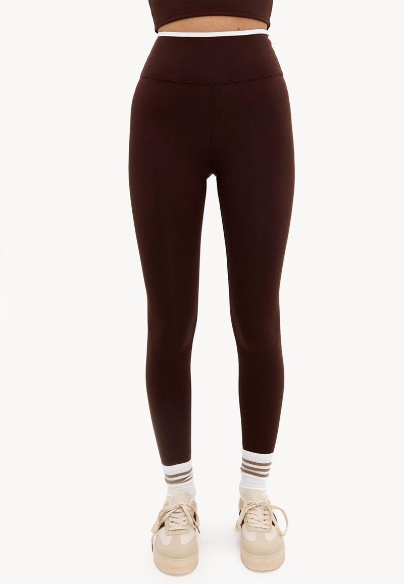 Personne portant des leggings marron foncé taille haute, des chaussettes blanches chevilles avec des rayures marron, et des baskets beige, debout sur un fond blanc uni.