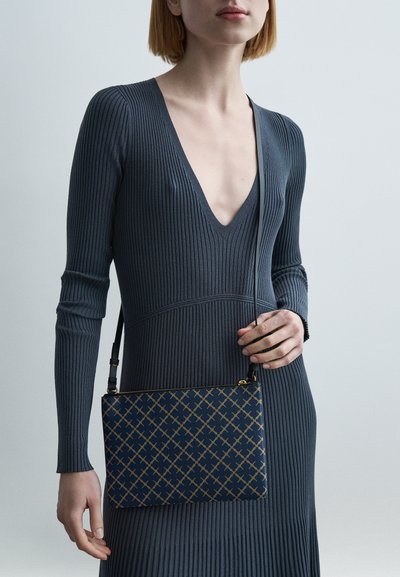 Sac à bandoulière bleu marine à motifs avec accents géométriques dorés, tissu texturé et bandoulière réglable. Porté avec une robe côtelée à col en V profond.