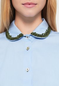 Lyseblå skjorte med knapper, der har en grøn, struktureret krave med fløjl-lignende finish og to knapper nær kraven.