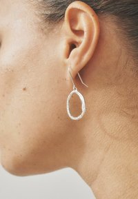 Pendientes de aro de plata con una superficie texturizada y un acento brillante, colgando de un gancho simple. La vista en primer plano muestra los detalles del diseño.