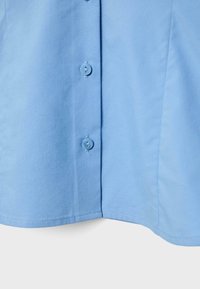 Jasnoniebieska tkanina z dwoma pionowo ułożonymi, przyszytymi guzikami w pobliżu środka, pokazująca dolną część koszuli.