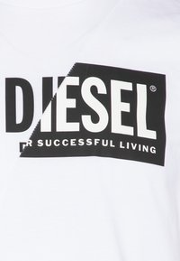 Valge T-särk musta tekstiga "DIESEL" ja "FOR SUCCESSFUL LIVING." Omab diagonaalset jagatud disaini ja siledat puuvillast tekstuuri.