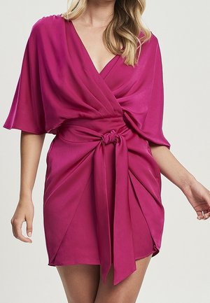 Kobieta ubrana w głęboko różową satynową sukienkę kopertową z szerokimi rękawami i przewiązaną w talii szarfą, stojąca na tle jednolitego jasnego tła.