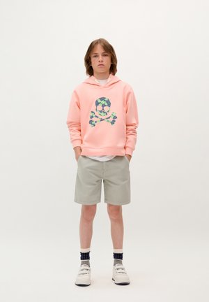 Sweat à capuche rose pâle avec un motif de crâne et de tibias bleus et verts, porté avec un short gris clair et des chaussures blanches avec des accents sombres.