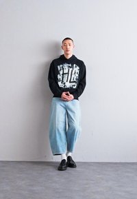 Sudadera negra con estampado gráfico blanco, jeans de pierna ancha de color azul claro y zapatos negros. El atuendo tiene un corte relajado y un estilo casual.