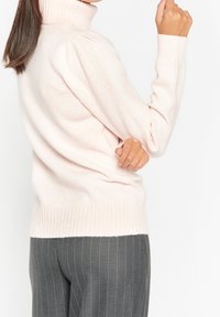 Pull-over en turtleneck rose clair avec des poignets et un ourlet à côtes, fabriqué dans un tissu doux et doté de manches bouffantes, associé à un pantalon à rayures grises.
