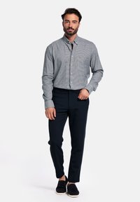 Man met een baard, gekleed in een grijs geruit overhemd met knopen, zwarte broek en zwarte instappers, staand tegen een effen witte achtergrond.