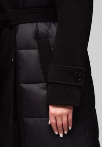 Hånd med hvide, polerede negle hviler ved siden af en sort, quiltet frakke med uldtekstur og knapdetalje ved ermet.