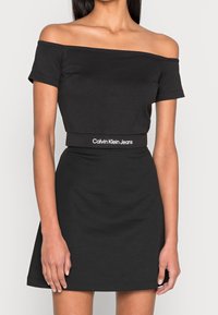 Vestido negro de hombros descubiertos con mangas cortas, cintura ajustada y falda acampanada. Presenta una banda de logo blanca en la cintura. Tela suave.