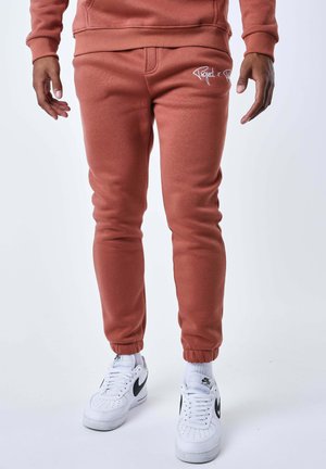 Hombre con pantalones jogger color óxido y zapatillas blancas Nike, de pie frente a un fondo liso.