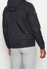 Schwarze Windjacke mit Kapuze, elastischen Bündchen und Saum. Mit einem Logo auf der linken Schulter. Hergestellt aus glattem, leichtem Material.