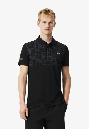 Polo nero con un motivo a griglia in diverse sfumature di blu nella parte superiore. Presenta un colletto e il logo verde Lacoste sul lato sinistro del petto.