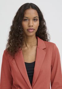 Blazer corail à finition texturée, à simple boutonnage et à revers crantés, porté sur un haut noir, avec une coupe ajustée et un tissu lisse.