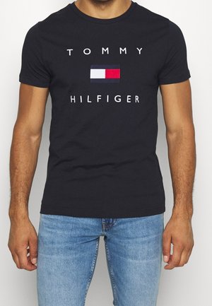 Mężczyzna ubrany w czarną koszulkę Tommy Hilfiger z białym, granatowym i czerwonym logo, w połączeniu z jasnoniebieskimi dżinsami, na jednolitym tle.