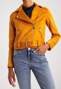 Gul lädermotorcykeljacka med midjebälte, asymmetrisk frontdragkedja och två sidofickor med dragkedja. Bärs över blå jeans.