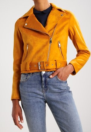 Giubbotto da motociclista giallo in pelle con vita cinturata, zip frontale asimmetrica e due tasche laterali con zip. Indossato sopra un paio di jeans blu.