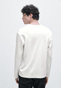 Langärmliges cremefarbenes Hemd aus weicher Baumwolle mit Rundhalsausschnitt, entspannter Passform und sauberen Nähten, kombiniert mit dunklen Jeanshosen.