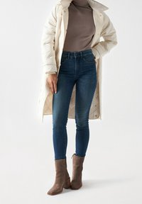 Abrigo largo beige acolchado, suéter de cuello alto ajustado marrón, jeans ajustados azul oscuro y botas de tobillo grises con tacón grueso y cremalleras laterales.