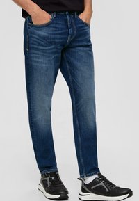 Dunkelblaue Denim-Jeans mit einem verwaschenen Muster, schmal zulaufenden Beinen, Fünf-Taschen-Design und kontrastierender Naht. Kombiniert mit schwarzen Sportschuhen.