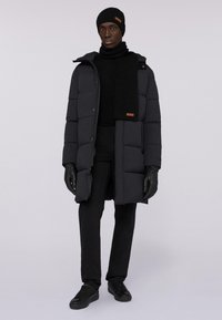 Cappotto puffa nero con trapping rettangolare, abbinato a una sciarpa nera, guanti e cappello. L'outfit presenta un design elegante e accenti minimi.