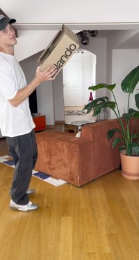 Jaunas vyras baltu marškinėliu ir juodu kepurėle laiko kartoninę dėžę moderniame svetainėje su rudais sofas ir dideliu žalio augalo.