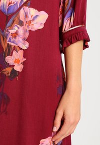 Bordeauxfarbenes Kleid mit übergroßen Blumenmustern in Rosa- und Lilatönen, ausgestattet mit einer Rüsche an den Ärmeln und einem dezenten metallischen Akzent.