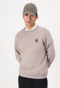 Világosszürke pulóver kerek nyakkal, vállán egy kis logóval. Egy megkopott szürke sapkával párosítva, amin szöveg található.