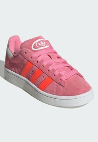 Sapatilhas de camurça rosa com três riscas laranja proeminentes. Apresentam atacadores rosa, sola de borracha branca e um logótipo na lingueta.