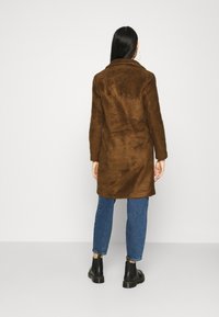 Hnědý kabát z umělé kožešiny s klopovým límcem, dlouhými rukávy a rovnými liniemi. Nesený přes modré džíny a černé kotníkové boty.