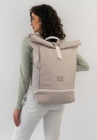 Beige roll-top backpack made of durable fabric, featuring a front pocket, white zipper detail, and a top handle for easy carrying.