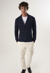 Tumesinine kootud kardigan shawl-krae, kolme nupuga, tekstureeritud kangas. Kombineeritud heledate pükste ja tumedate kingadega.