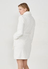 Accappatoio bianco in tessuto morbido con colletto a scialle, vita con cintura, tasche laterali e design di lunghezza a metà coscia. Texture liscia, decorazioni minime.