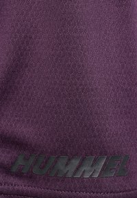 Lila strukturierter Stoff mit schwarzem „HUMMEL“-Markennamen nahe dem unteren Rand bedruckt.