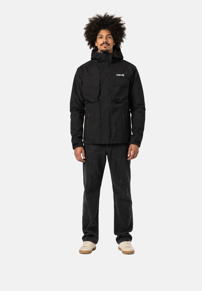 Mann mit lockigem Haar, der eine schwarze Kapuzenjacke, dunkle Jeans und weiße Sneakers trägt, steht frontal vor einem schlichten Hintergrund.