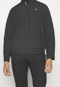 Black quilted jacket with a high collar, front zip closure, and logo detail on the left chest. Textured fabric with diamond patterns.