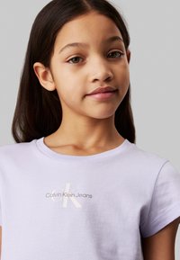 Camiseta de algodón lila con mangas cortas, que presenta un logo de "Calvin Klein Jeans" de color claro en la zona del pecho. Textura suave, corte clásico.