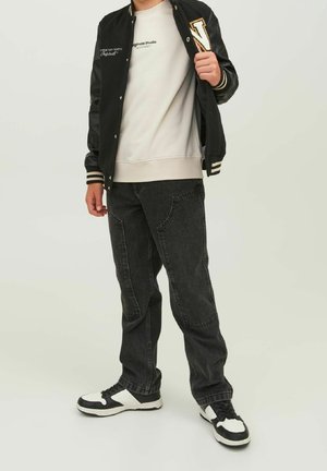 Person iført sort varsityjakke med bogstavet "N", hvid sweatshirt, sorte jeans og sort-hvide sneakers, der står mod en ensfarvet baggrund.