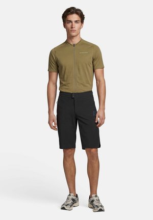 Junger Mann, der ein olivgrünes, zip-up Radsporttrikot, schwarze Radhosen und graue Sportschuhe trägt, vor weißem Hintergrund steht.