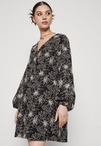 Robe noire avec broderie florale blanche, décolleté en V et manches longues bouffantes. Fabriquée en tissu léger avec une coupe ample.