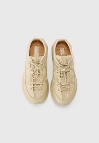 Beige sneakers med teksturert overdel, rund tå og polstret såle. Har snøringer og forsterket søm på sidene. Flat gummisåle.