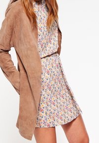 Südsbrun jacka som är lagd över en mönstrad klänning med ett diamantmönster i blått, gult och rött på en ljus bakgrund.