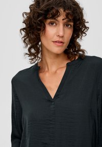 Schwarze, leichte Bluse mit V-Ausschnitt und strukturiertem Stoff. Mit einem kleinen Knopfdetail am Hals und langen Ärmeln.