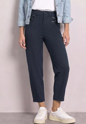 Femme portant un pantalon court bleu marine foncé avec des détails de boucle argentée, des baskets blanches, une chemise blanche et une veste en jean bleu clair aux manches retroussées.