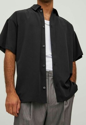 Homme portant une chemise noire à manches courtes boutonnée sur un débardeur blanc et un pantalon gris à rayures fines, avec une main dans la poche.