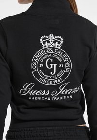 Giacca nera con un emblema ricamato bianco che recita "Los Angeles, California, Guess Jeans, American Tradition" e una corona sopra il testo.