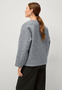 Maglione di lana grigio e strutturato con un ampio scollo rotondo e spalle scese; abbinato a pantaloni verdi scuri a vestibilità ampia.