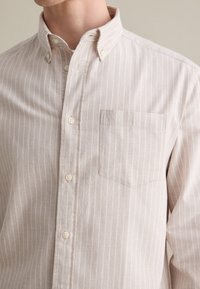 Camisa de botón beige claro con rayas verticales blancas, que presenta un collar pointed y un bolsillo en el pecho. Material de algodón con una textura suave.